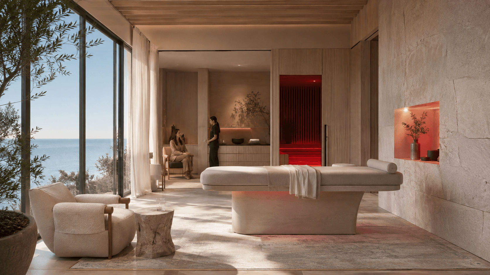 A calm treatment room with a therapy table, seating area and a glowing red-light sauna alcove, with two people conversing near a window overlooking the ocean.